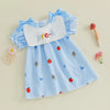 A baby girl's short-sleeve dress in light blue textured fabric with embroidered red strawberries, blue bows, and small fairy motifs. It features a white bib collar with the word 'pre-k' written in colorful letters, styled on a beige background with small decorative props.