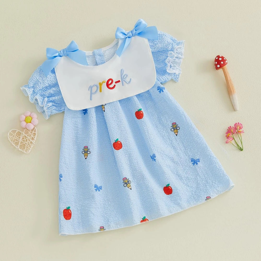 A baby girl's short-sleeve dress in light blue textured fabric with embroidered red strawberries, blue bows, and small fairy motifs. It features a white bib collar with the word 'pre-k' written in colorful letters, styled on a beige background with small decorative props.